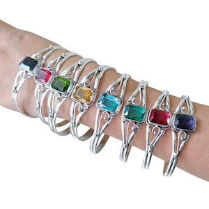 May include: A collection of eight silver-toned cuff bracelets, each featuring a single rectangular faceted gemstone in various colors including black, red, green, yellow, light blue, dark blue, pink, and purple, set in a decorative metal frame.