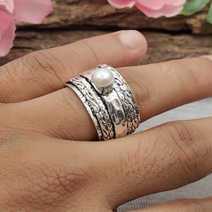 Può includere: Un ampio anello spinner in argento con una perla bianca centrale incastonata in una lunetta liscia. La fascia dell'anello presenta una finitura ossidata e testurizzata con intricati motivi floreali sui bordi e una sezione centrale in argento martellato.