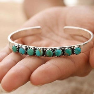 May include: A silver cuff bracelet featuring a row of eight round, polished turquoise stones. Each stone is set in a scalloped silver bezel, creating a continuous line of blue-green gems along the centre of the smooth, polished metal band.