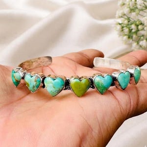 Könnte beinhalten: Ein silbernes Armband mit sieben herzförmigen Türkissteinen. Jeder Stein weist eine einzigartige natürliche Maserung in Blau- und Grüntönen auf und ist in einen polierten Silberrahmen gefasst, der sich der Form des Handgelenks anpasst.