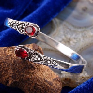 Puede incluir: Una pulsera de brazalete de plata con dos piedras preciosas rojas en forma de lágrima en los extremos abiertos. La banda de metal muestra intrincados grabados de volutas y patrones de cuentas decorativas alrededor de cada piedra.