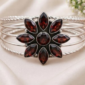 Könnte beinhalten: Ein Silberarmreif mit einer zentralen blumenförmigen Anordnung aus tiefroten, marquise-geschliffenen Granat-Edelsteinen. Das Armband besteht aus mehreren dünnen, gedrehten Silberdrähten, die von der zentralen Blumenfassung ausgehen.