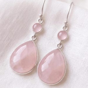 Peut inclure: Une paire de boucles d'oreilles pendantes avec des crochets et des montures en métal argenté. Chaque boucle présente une petite pierre ronde rose pâle au-dessus d'une pierre plus grande en forme de goutte, créant un design symétrique.