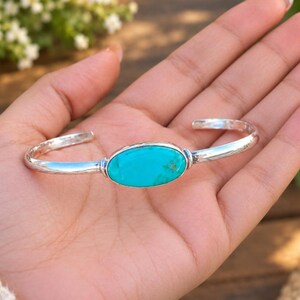 Könnte beinhalten: Ein silbernes Armband mit einem zentralen, ovalen Türkisstein. Das Metallband hat eine polierte, reflektierende Oberfläche und ein glattes, abgerundetes Profil. Der leuchtend blaue Stein ist horizontal in der Mitte des Armreifs gefasst.