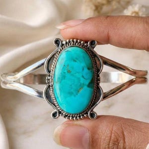 Könnte beinhalten: Ein Silberarmreif mit einem großen, ovalen Türkisstein in der Mitte. Der Stein ist von einer dekorativen gedrehten Drahtkante und einem gewellten Silberrahmen umgeben. Das Armband teilt sich an jeder Seite in zwei separate Silberarme auf.