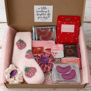 May include: A gift box containing a pair of fuzzy light pink socks with strawberry patterns, two satin scrunchies with purple tea lights, two sets of under-eye patches, a red strawberry-scented face mask, and a Monbana chocolate drink packet.