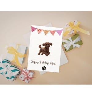 May include: A birthday card with a brown dog illustration, a paw print, and the text "Happy Birthday Max." The card is decorated with a banner of pink, orange, and purple triangles. Gift boxes with ribbons surround the card.