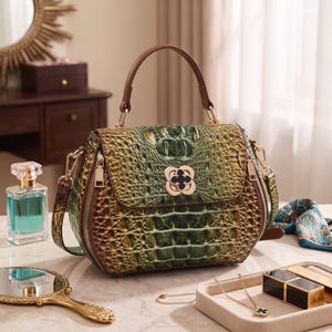 Puede incluir: Un bolso estructurado con textura de piel de cocodrilo en tonos verdes y marrones, que cuenta con un asa superior, un cierre dorado en forma de tr&eacute;bol en la solapa frontal y herrajes de metal dorado para una correa de hombro extra&iacute;ble.