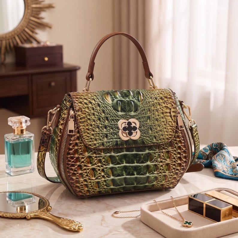 Puede incluir: Un bolso estructurado con textura de piel de cocodrilo en tonos verdes y marrones, que cuenta con un asa superior, un cierre dorado en forma de tr&eacute;bol en la solapa frontal y herrajes de metal dorado para una correa de hombro extra&iacute;ble.