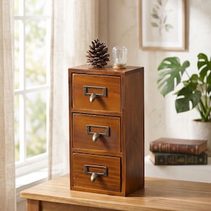 May include: A vertical wooden apothecary chest with three stacked drawers, each featuring a dark metal label holder and a curved pull handle. The chest has a medium brown wood grain finish and sits on a flat surface. Text reads HANDCRAFTED APOTHECARY CHEST.