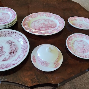 Può includere: Un set di stoviglie in ceramica bianca con un motivo a trasferimento rosa raffigurante paesaggi ed edifici. Il set comprende un grande piatto da portata rotondo, una pila di piatti più piccoli, un piatto ovale, una piccola ciotola ovale e un burriera con coperchio.