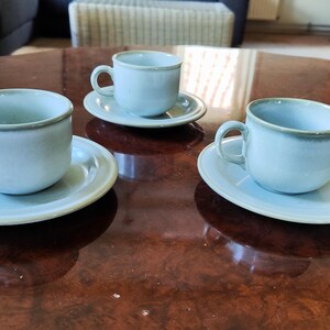 Op de afbeelding: Drie lichtblauwe keramische kopjes met bijpassende schoteltjes op een oppervlak. Elk kopje heeft een afgerond oortje en een gladde, effen afwerking met een iets donkerdere rand. De set bestaat uit drie individuele stuks drinkgerei.