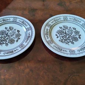 Może przedstawiać: Dwa okrągłe białe talerze ceramiczne ze szczegółowym czarnym wzorem kwiatowym pośrodku i pasującą ozdobną obwódką na krawędzi. Wzór składa się ze skomplikowanych ilustracji botanicznych przedstawiających różne kwiaty i liście.