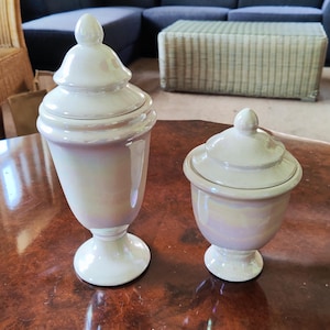 May include: Two white iridescent glass jars with matching lids, each featuring a pedestal base and a finial top. The taller jar has a slender, elongated body, while the shorter jar has a wider, more rounded shape, both displaying a pearlescent finish.