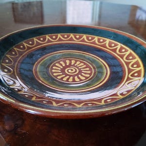 Könnte beinhalten: Ein runder Keramikt runder Keramikt runder Keramikteller mit glänzender Oberfläche und konzentrischen Ringen in Dunkelgrün, Braun und Creme. Die Mitte zeigt ein spiralförmiges Sonnenmotiv, während der äußere Rand mit einem wiederkehrenden Bogen- und Punktmuster verziert ist.