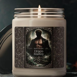 Puede incluir: Una vela perfumada blanca encendida en un tarro de cristal con tapa plateada. La etiqueta presenta la silueta de un hombre y el texto "Smells Like Draco Malfoy". La vela está sobre una superficie oscura.