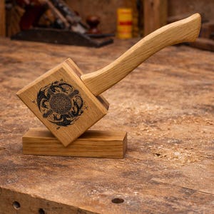 May include: A wooden mallet with a square head and a curved handle, resting on a rectangular base. The head features a black engraved design of dragons and Celtic knotwork. The mallet is made of light-coloured wood.