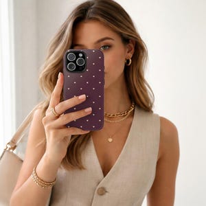 May include: A dark purple phone case with a pattern of small white hearts. The phone case is held up in front of the face. The person is wearing gold jewelry, including a necklace, earrings, and bracelets. The person is also wearing a beige vest.