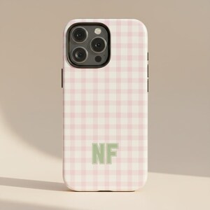 May include: A phone case featuring a pink and white gingham design. The case has a light green "NF" monogram in the centre. The phone case has a camera and volume buttons.