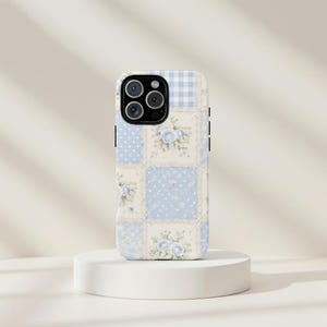 Peut inclure: Coque de téléphone avec un motif patchwork composé de carrés bleu clair à pois blancs, de motifs floraux et de vichy. L'étui a une bordure noire et une découpe pour l'appareil photo. L'esthétique générale est vintage et féminine.