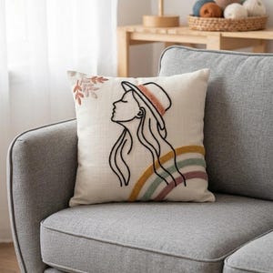 May include: Cream cushion with an embroidered design. It features a black outline of a woman in a hat, with rainbow and floral accents. The cushion is on a grey sofa. The sofa has wooden legs.