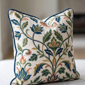 May include: A square scatter cushion with an off-white base featuring intricate embroidery of blue, green, and peach floral and vine patterns. The cushion is finished with dark blue twisted cord piping along the edges and a matching blue corner tassel.