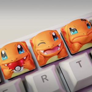 May include: Three orange Charmander-themed keycaps on a white keyboard. Each keycap features a different expression of the character. One holds a Pokéball, another is smiling, and the last is sad.