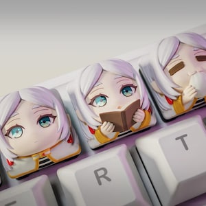 Op de afbeelding: Close-up van een mechanisch toetsenbord met aangepaste keycaps met anime-stijl personages. De keycaps zijn wit met gedetailleerde, kleurrijke illustraties van een meisje met wit haar en blauwe ogen. Eén keycap toont het personage dat een boek leest.