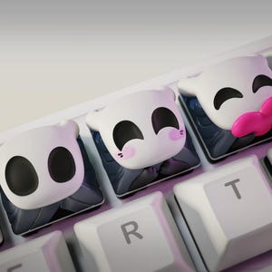 Puede incluir: Teclas personalizadas blancas y negras con caras de personajes de dibujos animados, incluida una con una lengua rosa, se muestran en un teclado blanco. Las teclas están en las teclas de las letras R, T e Y, y el número 3.