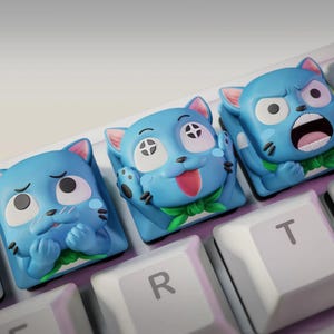 Pode incluir: Três teclas azuis em forma de gato com diferentes expressões faciais em um teclado branco. As teclas têm orelhas rosa e detalhes verdes, projetadas para teclados mecânicos. As teclas do teclado são brancas.
