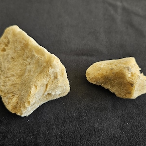 Pode incluir: Duas peças de cor bege claro do que parece ser pão ou um produto de panificação semelhante, sobre um fundo cinza escuro. As peças têm uma superfície áspera e texturizada.