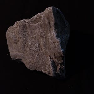 Può includere: Una roccia grigia ruvida con una superficie strutturata. La roccia ha un aspetto naturale e usurato, con variazioni di colore e consistenza. L'immagine è ambientata su uno sfondo scuro, evidenziando la forma della roccia.