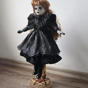 May include: A vintage-style doll in a black dress, with red hair and a pale face with dark eye makeup. The doll is mounted on a wooden stand. The dress has a patterned skirt and lace trim, creating a gothic aesthetic.