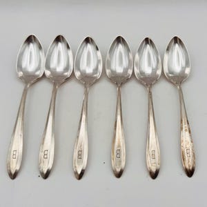 May include: A set of six silver-toned metal spoons arranged in a row. Each spoon features a pointed bowl and a tapered handle with a distinct letter B engraved near the base. The metal surfaces exhibit a polished finish with some visible tarnish.