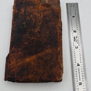 May include: A weathered, rectangular, brown leather-bound book with visible wear and tear. A stainless steel ruler with inch and centimeter markings is placed next to the book, showing measurements.