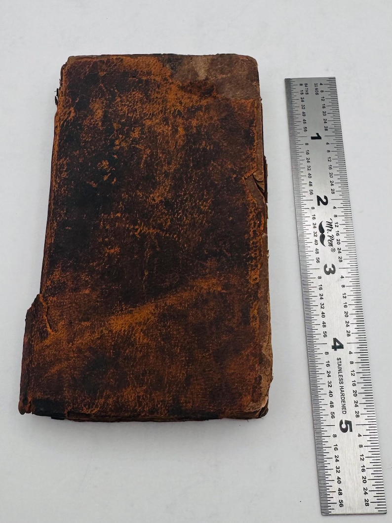 May include: A weathered, rectangular, brown leather-bound book with visible wear and tear. A stainless steel ruler with inch and centimeter markings is placed next to the book, showing measurements.