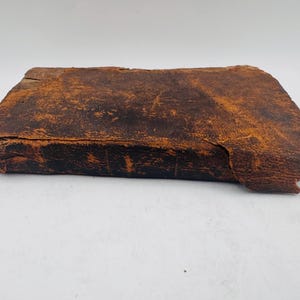 May include: A rectangular, antique leather-bound book with a distressed, weathered appearance. The cover is a deep brown with lighter orange-brown highlights, showing signs of age and wear. The edges are worn, and the overall aesthetic suggests a vintage item.