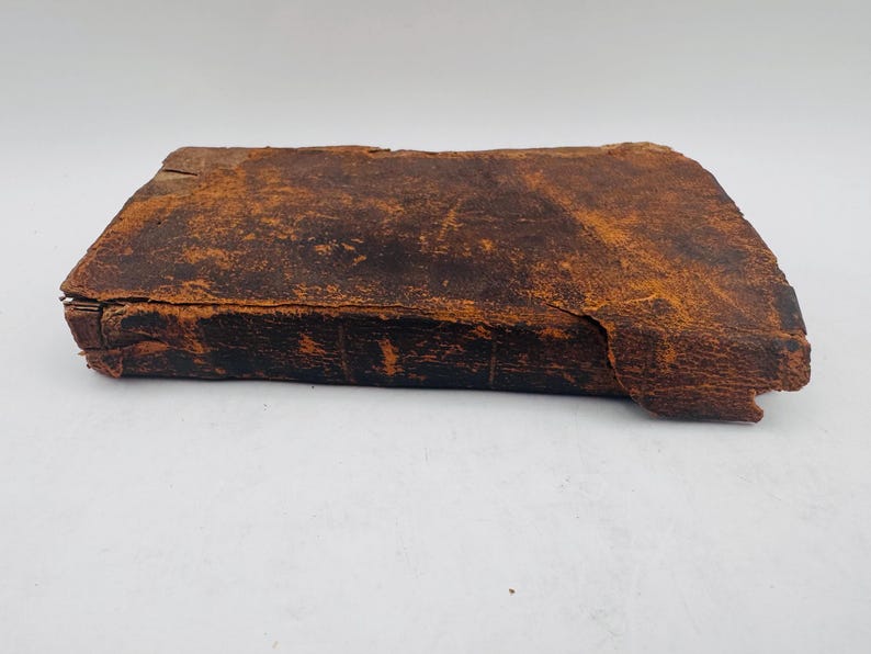 May include: A rectangular, antique leather-bound book with a distressed, weathered appearance. The cover is a deep brown with lighter orange-brown highlights, showing signs of age and wear. The edges are worn, and the overall aesthetic suggests a vintage item.