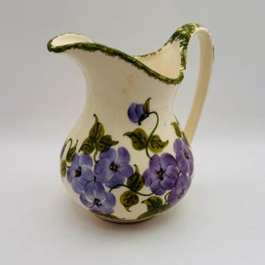 May include: Cream-colored ceramic pitcher with a handle and a spout. The pitcher is decorated with hand-painted purple flowers and green leaves. The rim and handle are trimmed with green paint. A decorative floral pitcher.