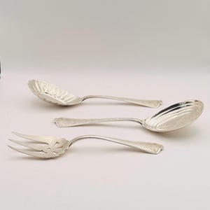 May include: Three pieces of silver serving cutlery: a slotted spoon, a serving spoon, and a serving fork. The spoon bowls have decorative edges and the handles have a floral design. The pieces are arranged on a white background.
