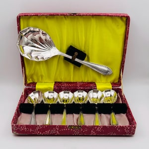 May include: A silver-toned serving spoon and six matching smaller spoons, each featuring a shell-shaped bowl. The set is presented in a rectangular case with a bright yellow satin interior lining and a textured, patterned red exterior cover.