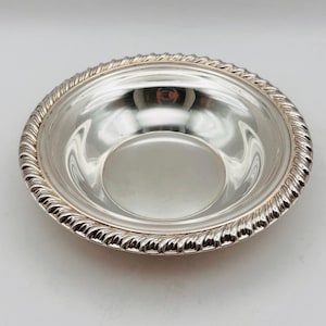 May include: A round, polished silver bowl featuring a reflective, smooth interior surface and a decorative, raised rope-style border along the outer rim. The metallic finish creates clear reflections of the surrounding environment on its surface.