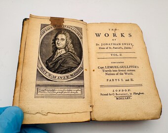 May include: An open antique book with aged, yellowed pages. The left page features a portrait of Captain Lemuel Gulliver, while the right page displays the title "The Works of Dr. Jonathan Swift." The book is bound in dark brown leather.