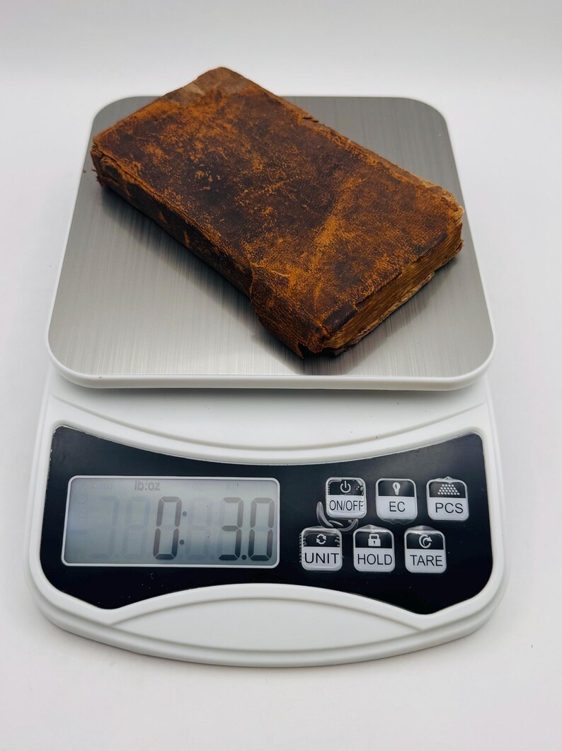 May include: A small, antique book with a worn, brown leather cover is placed on a digital scale. The scale displays a weight of 3.0 oz. The book's pages are visible, and the overall aesthetic suggests age and use.
