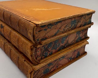 May include: Three antique books stacked on top of each other. The books have tan leather covers with gold trim and marbled endpapers in shades of red, blue, and black. The edges of the pages are gilded.