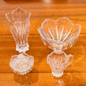 May include: Four-piece clear crystal glassware set. Includes a tall vase, a serving bowl, a small lidded dish, and a creamer. The pieces have a cut glass design and are arranged on a wooden surface.