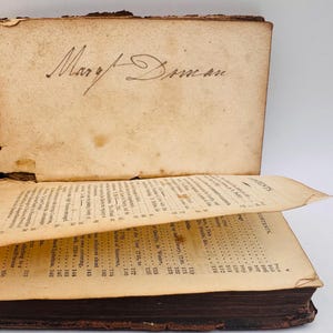 May include: An antique book with aged, yellowed pages. The cover is open, revealing handwritten script that reads "Mary Duman." The pages display printed text and a table of contents. The book's binding is dark brown.