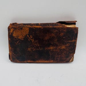 May include: A rectangular, antique book with a worn, brown leather cover. The leather shows signs of age with dark brown and tan patches. The top edge of the book is damaged, revealing the aged pages within. The book is set against a white background.