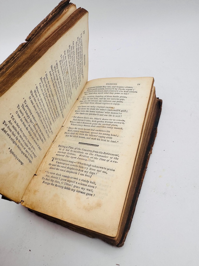 May include: An open antique book with aged, yellowed pages. The book's cover and edges show signs of wear, with a visible spine. The text is in English, with the title "Elegies" visible on one page. The book is set against a white background.