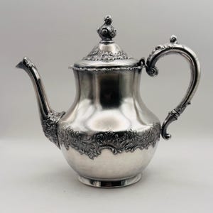 May include: A silver teapot with a curved spout and handle. The teapot has a lid with a decorative finial and ornate floral detailing around the base and lid. The teapot is made of a shiny, reflective metal.